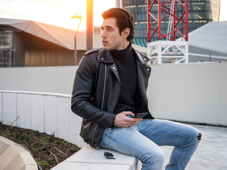 One handsome young man in urban setting in modern city, sitting, wearing black leather jacket and jeansの写真素材