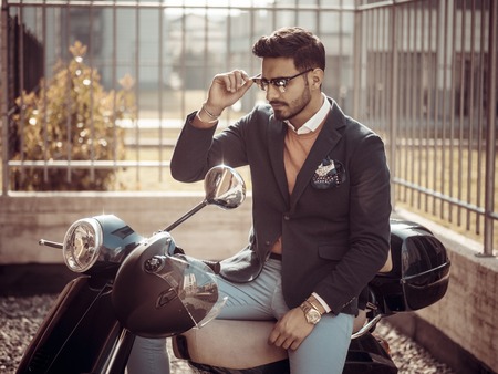 Young stylish man in suit and sunglasses standing by his classic Italian scooter on empty road in sunny city.の写真素材