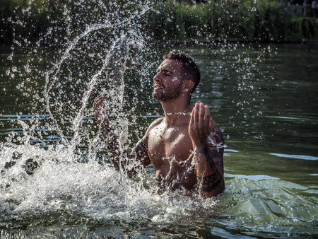 Handsome, hot young bodybuilder in the sea, splashing water up, showing his muscular torso and armsの写真素材
