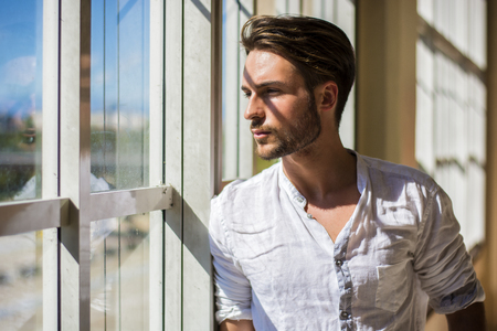 Handsome serious young man standing inside modern building next to big window, wearing white shirt, looking outの写真素材