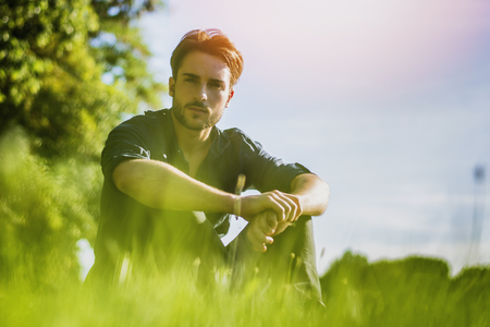 Attractive, fit young man relaxing sitting on lawn in the countryside among grass, looking at cameraの写真素材