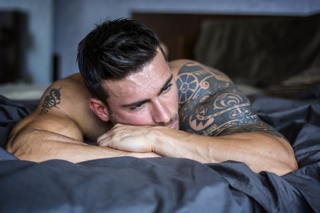Shirtless muscular sexy male model lying alone on bed in his bedroom, looking away with a seductive attitudeの写真素材