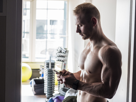 Shirtless muscular blond young man using cell phone in gymの写真素材