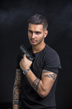 Young handsome man holding a hand gun, wearing black t-shirt, on black background in studioの写真素材