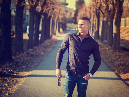 One handsome young man in urban setting in European city walking along street with trees, wearing black jacket and jeansの写真素材