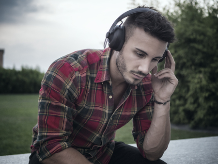 Young attractive man in city setting listening to music with headphones, sitting on benchの写真素材