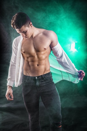 Handsome young muscular man shirtless wearing jeans, taking off leather jacket on naked muscle torso, on dark background in studio shotの写真素材