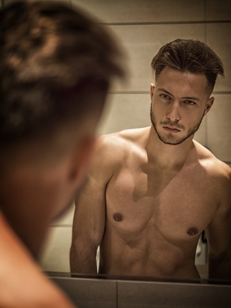 Shirtless muscular handsome young man looking at himself in bathroom mirror in the morningの写真素材