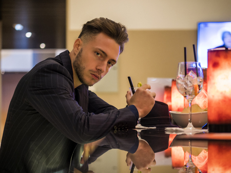 Handsome young man drinking cocktail at bar counter, wearing business suit and looking at cameraの写真素材