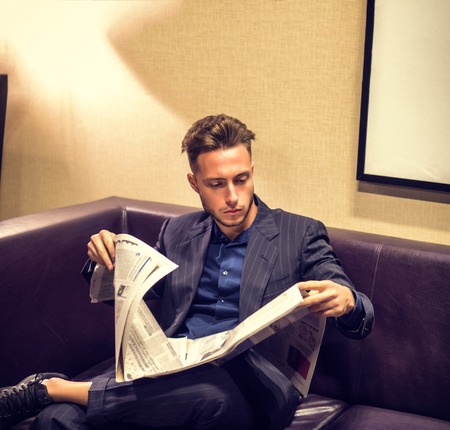 Handsome young man at home reading newspaper, sitting on a leather sofaの写真素材