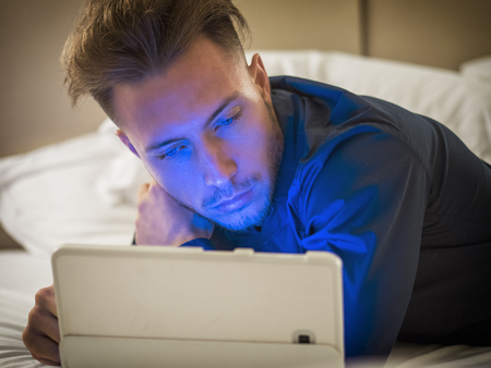 Handsome young man in bed typing on tablet PC, sending text message or dialing number or reading web pagesの写真素材
