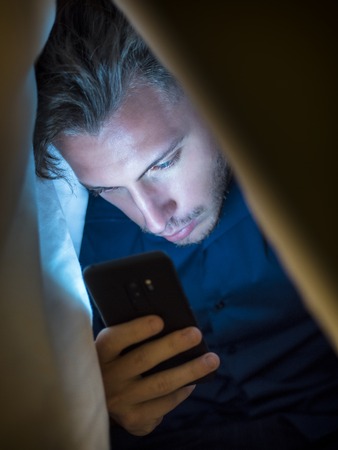 Young man hiding himself under a blanket while using a mobile phone for browsing the internet or for chatting online privatelyの写真素材