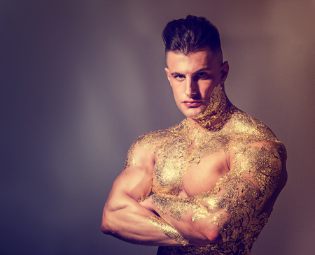 Muscular naked man covered with gold, with arms crossed on chest and looking sensually at camera on black backgroundの写真素材