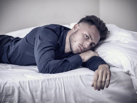 Sexy male model laying alone on bed in his bedroom, looking at camera with a seductive attitude,, wearing blue elegant shirtの写真素材