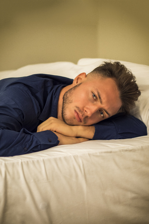 Sexy male model laying alone on bed in his bedroom, looking at camera with a seductive attitude,, wearing blue elegant shirtの写真素材