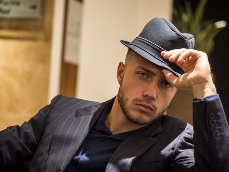 Handsome elegant man in hotel or night club lounge, wearing business suit and fedora hat, looking at...