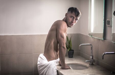 Shirtless young man in bathroom looking at camera, by sink and mirror, in the morningの写真素材