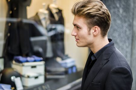 Handsome Young Man in Black Elegant Suit Looking at Displayed Fashion Items in Glass Window Boutique at the Street Side.の写真素材