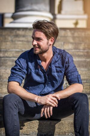 One handsome young man in urban setting in summer day, wearing blue shirt, sitting on stone steps