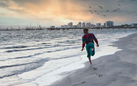 Back view of anonymous barefoot male with dreadlocks running on wet sand near waving sea against coastal city and cloudy sundown sky with birdsの写真素材