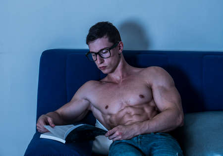 Young male bodybuilder reading book sitting on sofaの写真素材