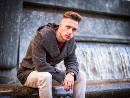 Young man sitting by fountain, looking at camera in city setting