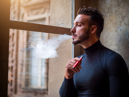 Handsome man vaping, smoking e-cigarette by a windowの写真素材