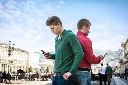A couple of men standing next to each other, back to back, engrossed in their cell phones and...