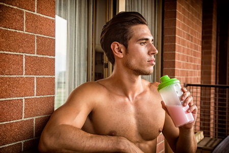young and attractive muscular shirtless man enjoying a colorful protein shake, sitting outdoor next to a brick wallの写真素材