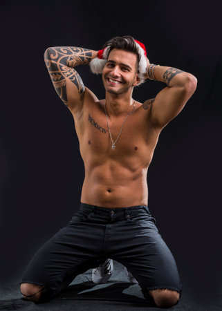 A shirtless man with a santa claus hat on. Photo of a shirtless man wearing a Santa Claus hat in studio, kneeling on the ground on black backgroundの写真素材