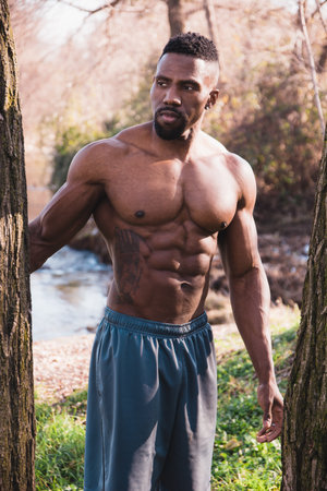 A shirtless man standing next to a tree. Photo of a shirtless man standing next to a tree in a...