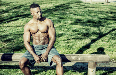 A shirtless muscular black man sitting on a bench in a park. Photo of a shirtless man enjoying nature in a peaceful parkの写真素材