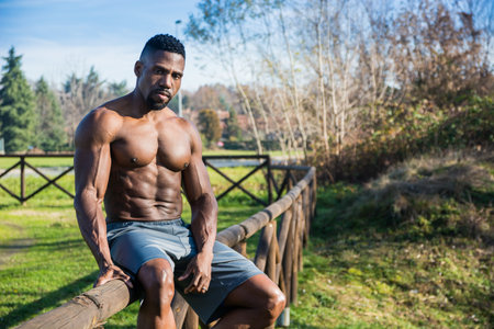 A shirtless man leaning on a wooden fence. Photo of a muscular African American man leaning on a...