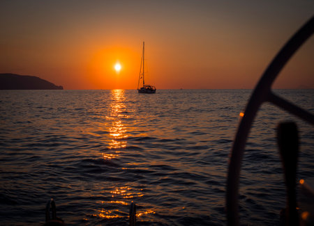 The sun is setting over the ocean with a sailboat in the distance and a helm in the foreground. Photo of a colorful sunset over the ocean with a sailboat in the distanceの写真素材