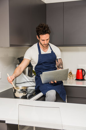 A man sitting on a kitchen counter using a laptop, reading a recipe while stirring a pot. A Handsome, Muscular Man Sitting on a Kitchen Counter with a Laptopの写真素材