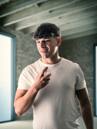 A young man in a white t - shirt is spraying cologne or perfume on himself, smilingの写真素材