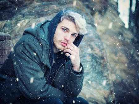 Portrait of young man in hoodie posing outdoor in winter setting with snow all around, looking at cameraの写真素材