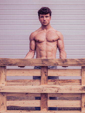 A young man with a muscular build stands proudly behind a wooden pallet. His bare torso highlights his fitness. The setting has a neutral background that emphasizes his presence.の写真素材