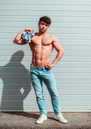 A muscular young man stands confidently outside, holding a towel over his shoulder. He wears jeans and white sneakers, showcasing a fit physique in the bright sunlight.の写真素材