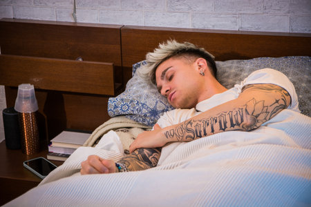 A young man with blond hair and blue eyes is lying in bed, relaxed and contemplative. His tattoos are visible as he rests, surrounded by a cozy atmosphere in the morning light.の写真素材