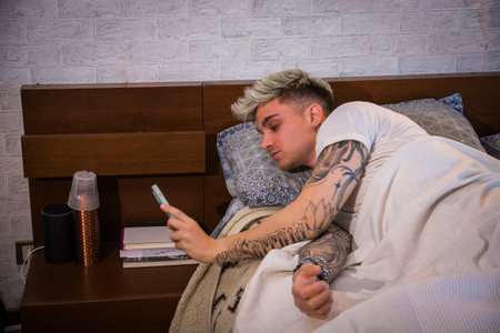 A young man with stylish blond hair and tattoos is comfortably lying in bed, focused on his phone. The soft light of morning fills the cozy bedroom, enhancing the relaxed atmosphere.の写真素材