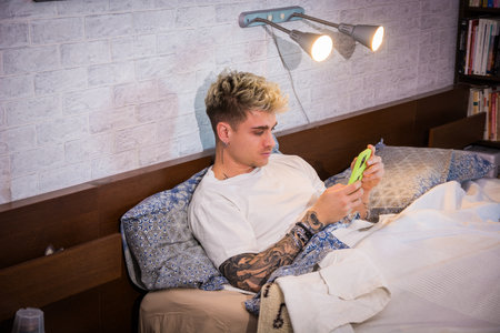 A young man with blonde hair and blue eyes sits comfortably in bed, focused on his phone. The cozy...