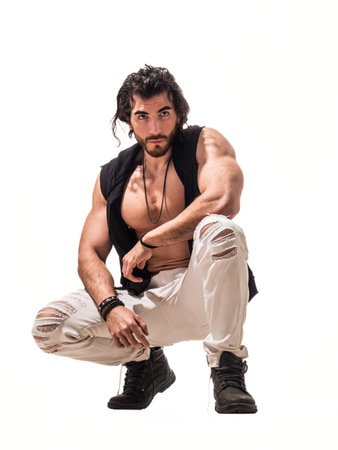 Muscular man crouches in a striking pose, showcasing a stylish, casual outfit featuring ripped jeans...