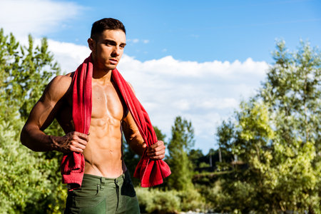 A male model stands confidently by a river, showcasing his physique while holding a red towel. The...