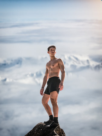 A young man with athletic build stands confidently on a rocky mountain peak, surrounded by...