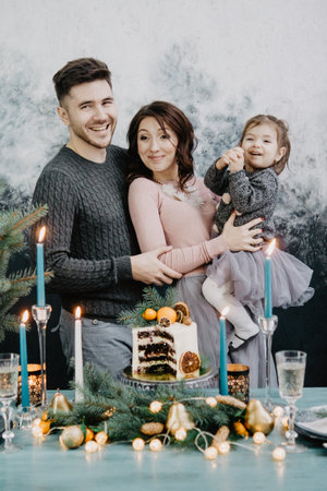 Young family with a daughter in festive outfits at a served table with candles, garlands, sparklers and a cake near the Christmas treeの写真素材