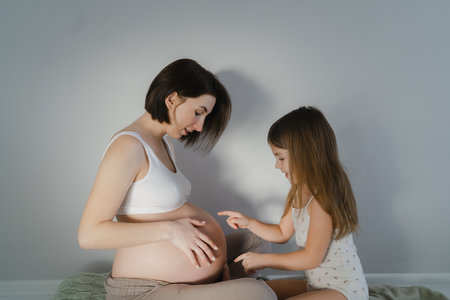 Young pregnant girl with a little daughter in a bright room in white home clothesの写真素材