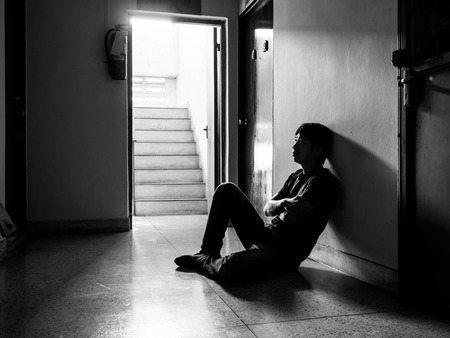 a sad young man sitting in the dark leaning against the wall with his back, The sun is in sorrow, black and white.の写真素材