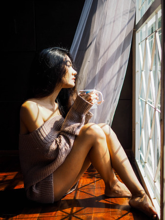 Young beautiful woman with cup of coffee sitting down by the window.の写真素材