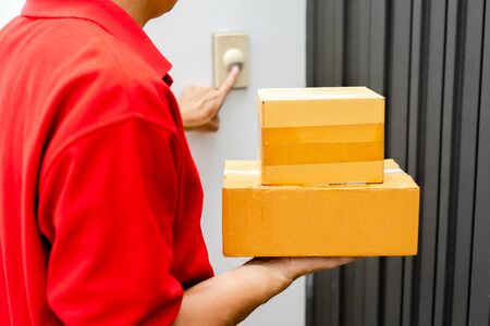 Delivery man in red uniform standing at the door with parcel post box press ring bell in front of the house to delivery of the good, Deliver packages to recipients quickly, Home delivery concept.の写真素材